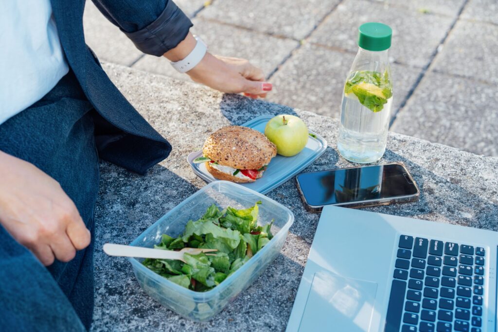 Una chica come ensalada y un bocadillo mientras usa su portátil
